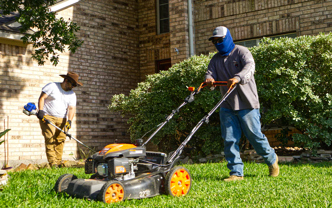 Two people mowing and trimming a residential lawn, working opposite ends of the same yard.