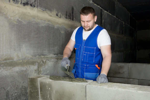 A mason in a blue work vest setting a concrete block into a course with a trowel.